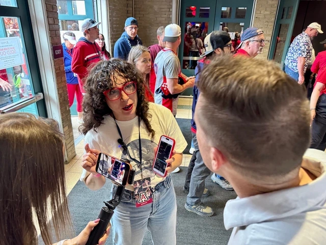 A woman with curly hair and red glasses, holding a phone and talking to a man, surrounded by a crowd in casual attire. The scene is lively and engaged.