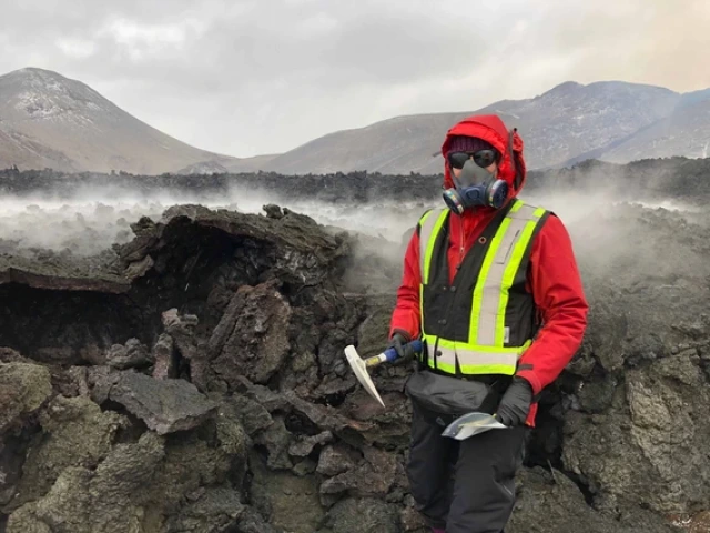 Wearing an orange safety vest and a gas mask, Solange Duhamel stands next to a wall of jumbled lava rocks reaching up to her shoulder. 