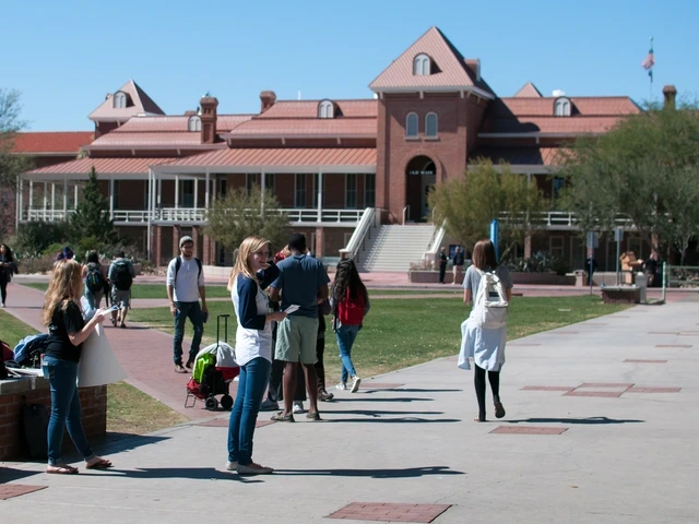 Students walk and gather on a campus mall in front of Old Main, a red-brick building with a wraparound porch and central staircase, on a sunny day.