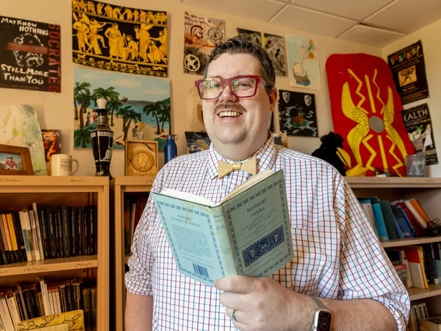 A man in a white and red patterned buttondown and red glasses stands smiling in front of a bookcase while holding a light blue copy of The Odyssey