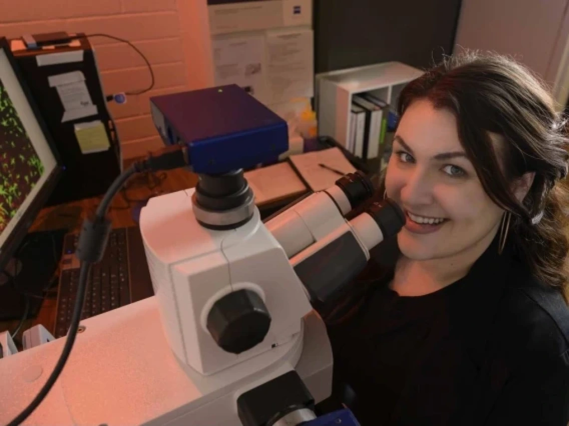 This image shows a woman smiling while working in a laboratory setting, looking into the eyepiece of a microscope. A computer screen in the background displays a vibrant, microscopic image, likely of a biological sample, alongside laboratory equipment and notes on the desk.