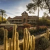 A historic brick building stands behind a garden filled with blooming red and purple flowers, tall cacti, and a fountain spraying arcs of water. Palm trees frame the scene as warm late-afternoon sunlight casts long shadows across the walkway.