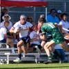 A rugby match with two male players in action. The player on the left, in a white jersey with blue and red accents, holds a rugby ball and is sprinting forward. He wears a protective headband with a star pattern. The player on the right, wearing a green jersey marked with the number 11, is approaching to tackle. Both players are on a grassy field with sunlight casting shadows.