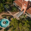 Aerial view of a historic red-brick building with a brown metal roof and a prominent staircase leading to the entrance. In front of the building, an octagonal water fountain with a central pedestal is surrounded by paved walkways, manicured gardens, and green trees. The scene is bathed in warm sunlight, casting long shadows across the landscape.