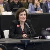 A woman in a black blazer speaks into a microphone while seated at a table during a formal meeting. She holds a small device in one hand, with an open laptop displaying several stickers in front of her. People sit in rows behind her, listening attentively.