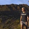 A man wearing shorts and a black t-shirt that says "ARIZONA" is hiking in a desert landscape, with a mountain in the background.